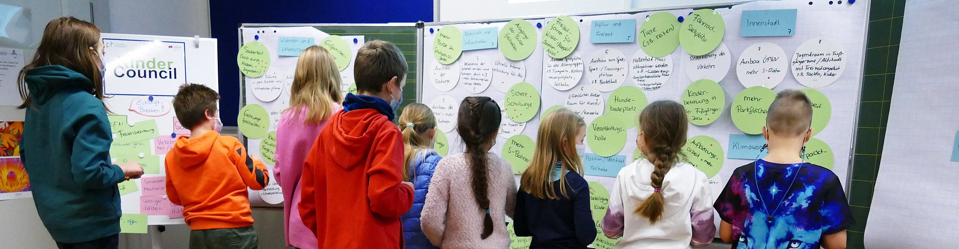 Mehrere Kinder stehen vor einer Tafel mit angehefteten Kreisen aus Papier, auf die sie ihre Ideen aufgeschrieben haben. Die Ideen werden gerade geclustert und priorisiert.