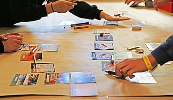 Eine mit Packpapier belegte Tischplatte, auf der verschiedene Hände und Arme zu sehen sind, die mit den Puzzleteilen gerade das Serious Game "Klimapussle" spielen
