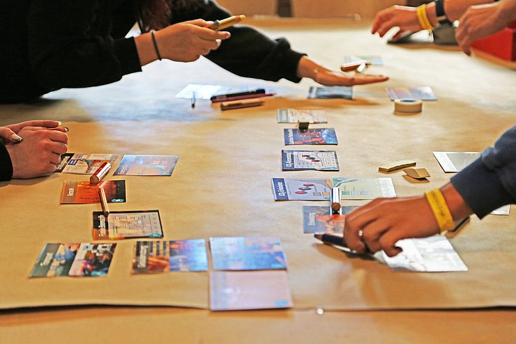 Eine mit Packpapier belegte Tischplatte, auf der verschiedene Hände und Arme zu sehen sind, die mit den Puzzleteilen gerade das Serious Game "Klimapussle" spielen